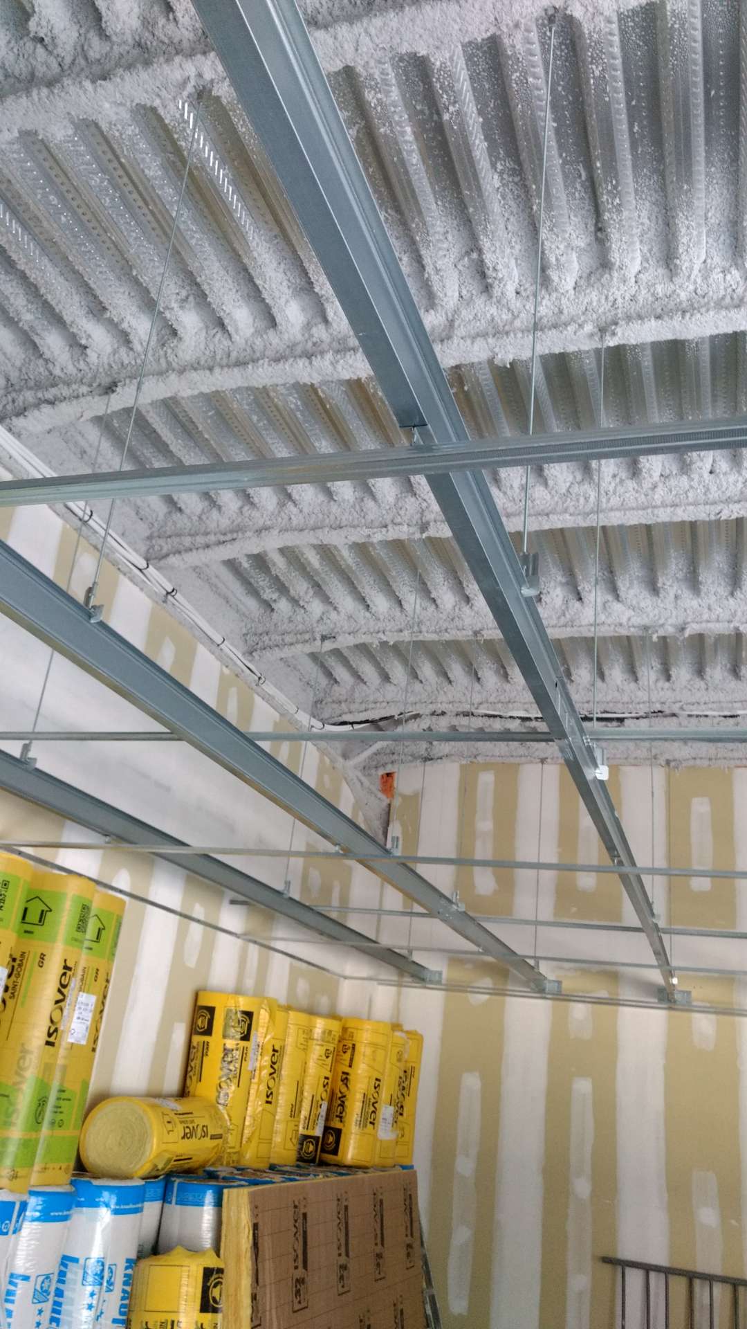 Plafond métallique nervuré recouvert de mousse isolante blanche, avec ossature suspendue et rouleaux d'isolant jaune empilés contre un mur en plaques de plâtre.