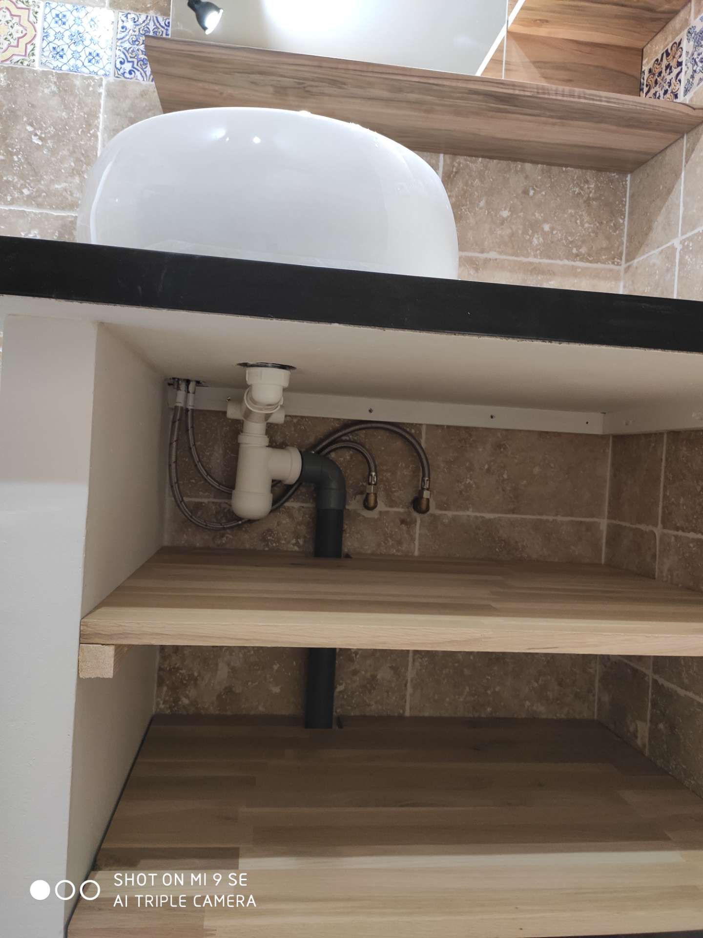Sous un lavabo blanc, une étagère en bois laisse passer la tuyauterie d'évacuation et deux flexibles, dans un meuble carrelé beige.