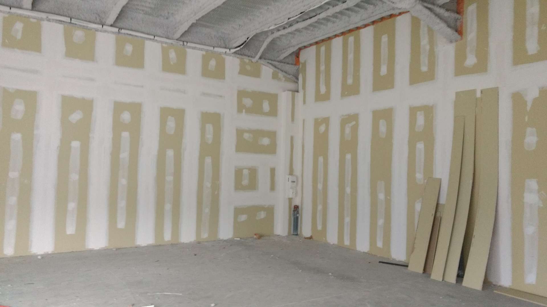 Pièce en travaux aux murs de plaques de plâtre jointoyées, sol et plafond bruts, plaques posées contre le mur de droite.