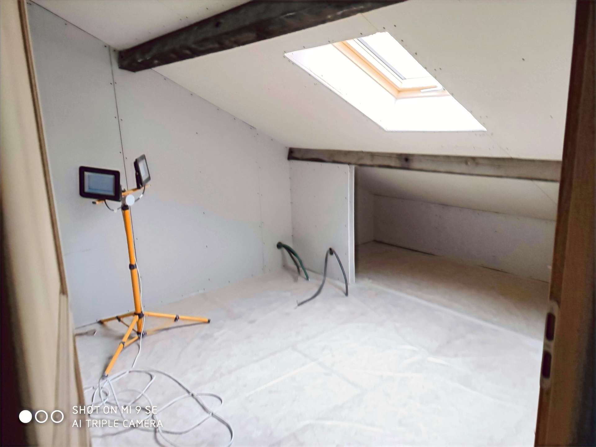 Combles en travaux : sol et murs couverts de plaques blanches, poutre apparente, fenêtre de toit, projecteur sur pied jaune et tuyaux au sol.